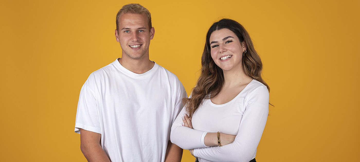 Downloads für Einsatzstellen. Zwei Personen in weißen T-Shirts stehen vor einer gelben Wand. Beide schauen und lächeln in die Kamera.